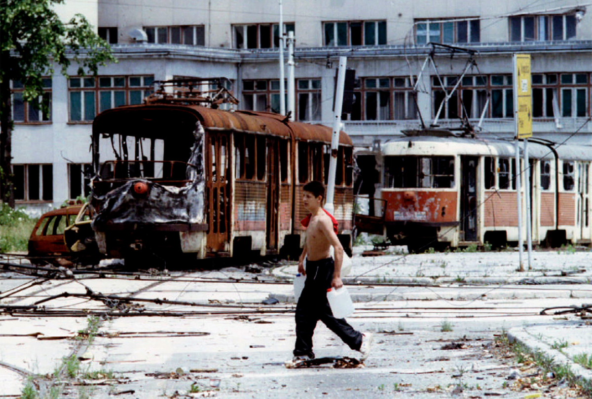 (пред) Тинејџер носи вода пред уништените трамваи на плоштадот Скендерија на 22-ри јуни 1993.